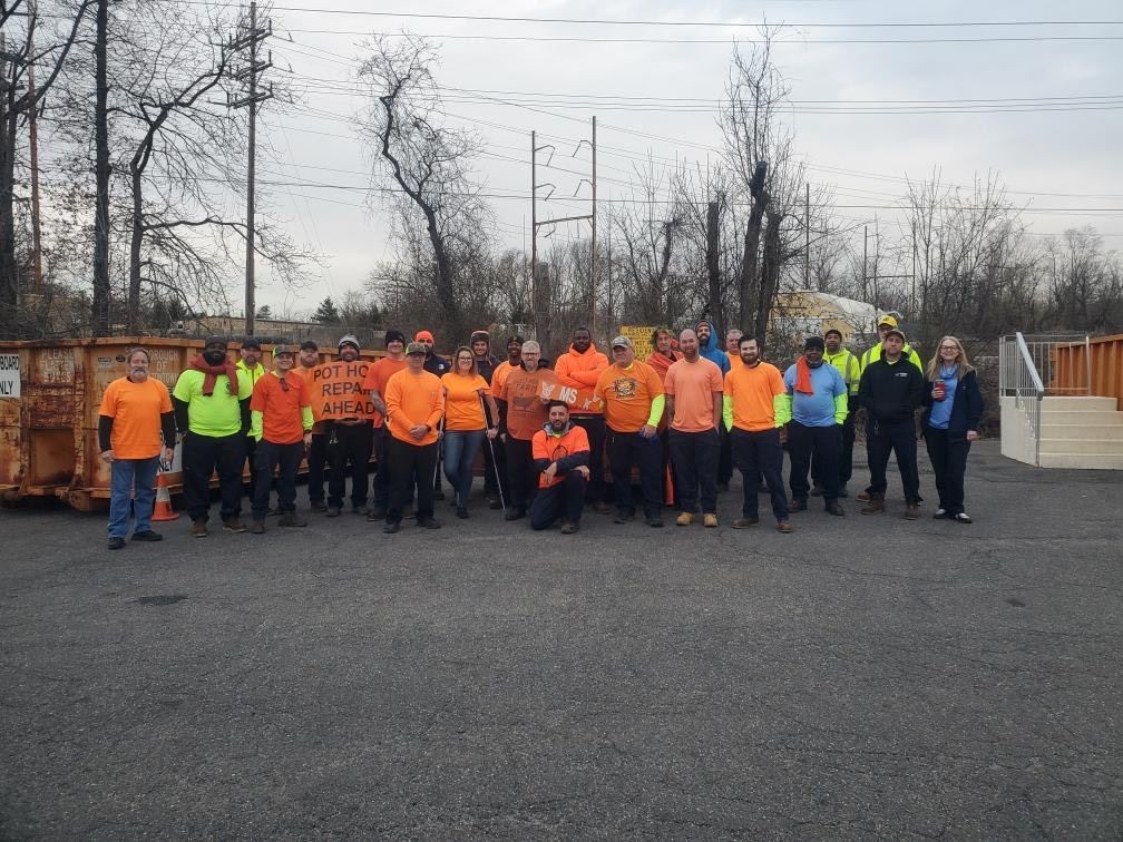 Group of volunteers in orange and neon shirts posing outdoors on a paved lot near construction equipment.